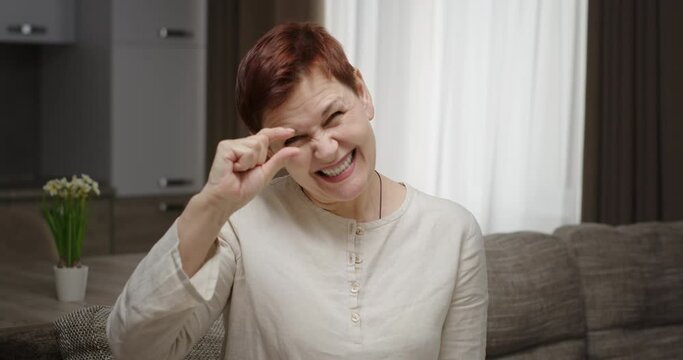 Funny woman asking for favor. Mature female with short hair gesticulating and looking at camera while sitting on sofa and pleading for small favor at home