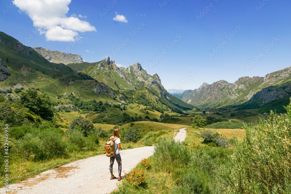 Wall mural A girl with a backpack is looking to the mountains and there is a blue sky and some white clouds