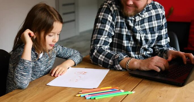 Father working in his home office on a laptop, her daughter sits next to her and draw, Man freelance, remote work and raising a child at workplace.