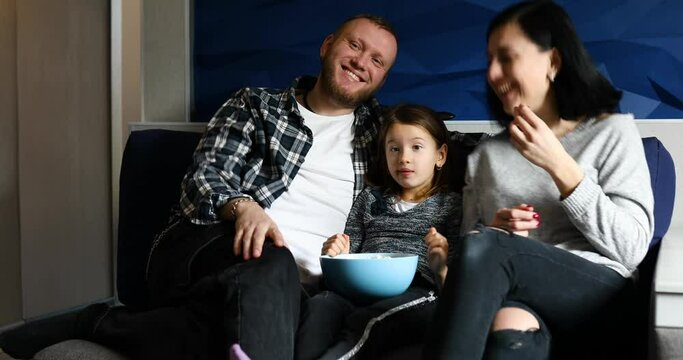Mother, father and daughter eat popcorn and watch TV film, watching a movie on a couch at home, happy smiling family spending free time together, relax.