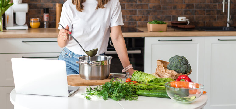 Close up woman in white t-shirt cooking soup with fresh vegetables in kitchen at home. Menu, recipe book banner. Girl reads the recipe in laptop. Caucasian model using internet in loft apartment.