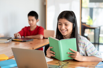 Obraz premium Image of a happy Asian girl wearing headphones holding a textbook online learning using a laptop in the classroom, the background is blurred.