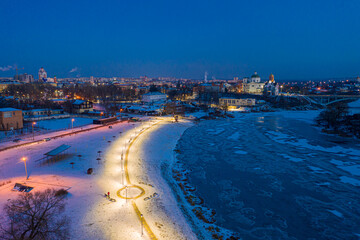 Naklejka na ściany i meble Beautiful evening top view of the city. Evening, night illumination in the city. Winter city in the snow. The river is covered with ice.