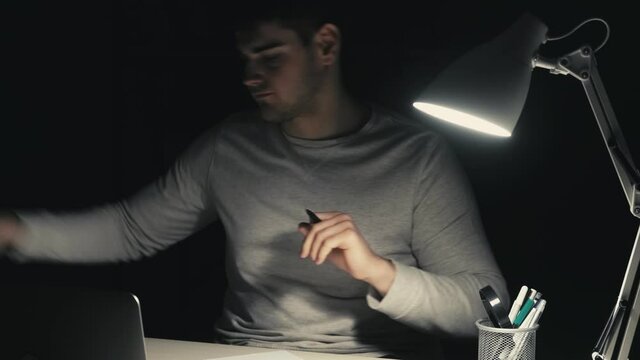 Overwork burnout. Night stress. Creative block. Late work anxiety. Tired annoyed sleepy left-handed man dissatisfied with writing ideas creasing throwing away paper at dark home workplace.