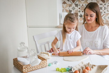 Happy mom and daughter prepare dough together using rolling pin, smiling young mom, charming girl prepare cookies, buns in the kitchen. The concept of a friendly family preparing for Easter