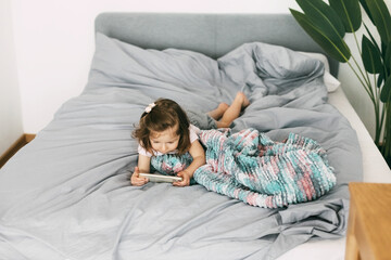 A little girl watches cartoons on her phone or plays on the bed before going to bed. Top view