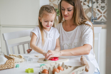 Happy mom and daughter prepare dough together using rolling pin, smiling young mom, charming girl prepare cookies, buns in the kitchen. The concept of a friendly family preparing for Easter