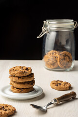 Top view of chocolate cookies on white plate, spoon and glass jar with cookies, on white table, black background, in vertical