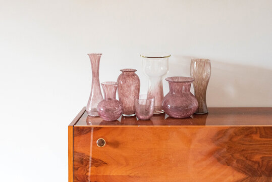 Mid-century purple pink modern artisan glass vase collection on a wooden table with plants before the window