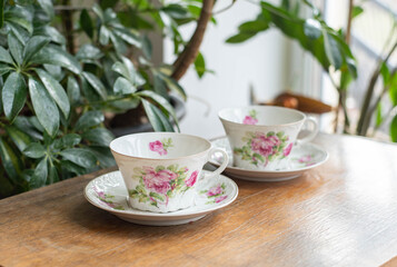 Vintage porcelain coffe cup set with rose pattern on a wooden table before teh window with plants