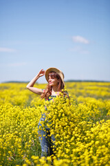 Naklejka na ściany i meble Young, beautiful woman in rapeseed field in the summer. Rural scene with attractive girl in hat enjoying sun in yellow blooming field. Concept of joy, happiness and freedom.
