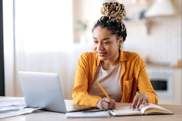 Fototapeta Focused cute stylish african american female student with afro dreadlocks, studying remotely from home, using a laptop, taking notes on notepad during online lesson, e-learning concept, smiling obraz