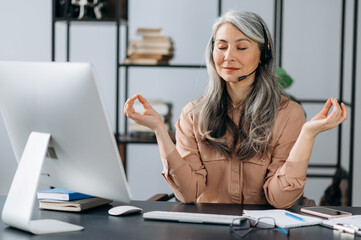 Obraz na płótnie Canvas Relaxed calm beautiful mature gray-haired asian woman, business leader, call center operator, in headset, in stylish clothes, resting at workplace, meditation, relieve stress by closing her eyes