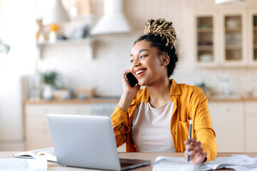 African American joyful attractive stylish young woman, freelancer, manager or real estate agent, having pleasant phone conversation with client or employee, sitting at workplace, smiling - obrazy, fototapety, plakaty