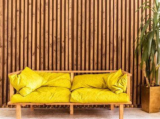 A trendy yellow sofa stands against a background of a wall with wooden slats. A large flowerpot with a plant stands next to the sofa