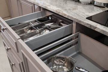 Opened drawers with cutlery, utensils, pots and pans inside in beige kitchen with marble countertop