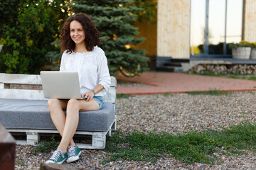 Smiling curly young woman dressed in casual clothes, working in her yard at home on a laptop.