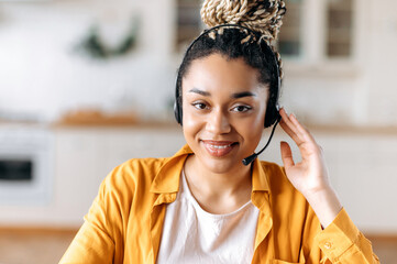 Close-up of African American an attractive confident young woman in headset, sitting in office, working as operator of call center or support service, looks directly at the camera and smiling friendly