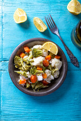 Broccoli, cauliflower and carrot salad in bowl on blue wooden table with olive oil and lemon slices. Top view.