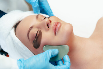 Beautiful woman having guasha scraper face massage done by cosmetologist