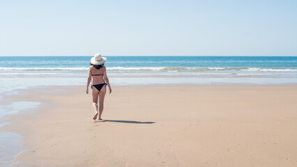 Young brunette woman with curly hair wearing black bikini and hat walking along a lonely beach. vacation, leisure, relaxation and tranquility concept