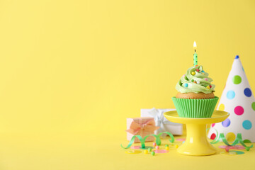 Delicious birthday cupcake with burning candle, party decor and gift boxes on yellow background, space for text