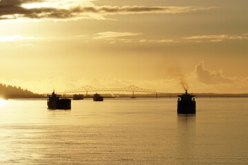 Obraz premium View from container ship on the other merchant cargo vessels anchored near to entrance to Columbia river from Pacific ocean where is situated port of Portland during sunset with bridge in background.