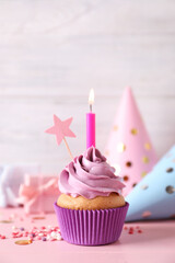 Birthday cupcake with burning candle, party hats and gift boxes on pink table