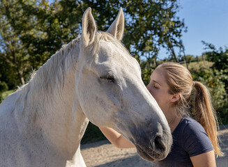 Pferdeliebe: junge blonde Frau und ihr Schimmel schmusen mit geschlossenen Augen auf dem Reitplatz in der Sonne