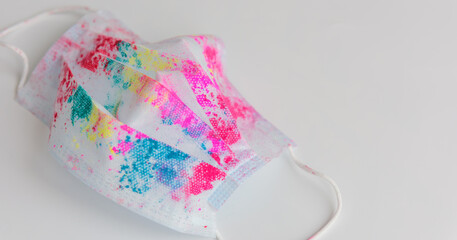Used medical face mask with bright multicolored spots from holi paints, on white background, close-up. Concept of celebrating the Indian Happy Holi Festival during coronavirus pandemic. Copy space.