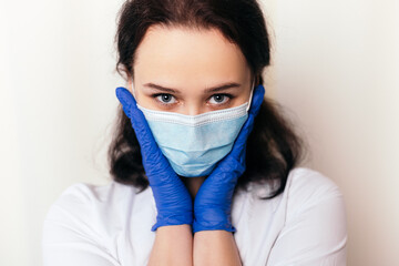 Nurse shy young medic puts on rubber disinfected gloves. Female doctor dressed in medical gown and surgical face blue mask. Image with copy space. Pandemic virus coronavirus concept

