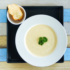Beige chicken cheese cream soup with toasted white bread and one herb in the middle of plate. White and blue wooden surface