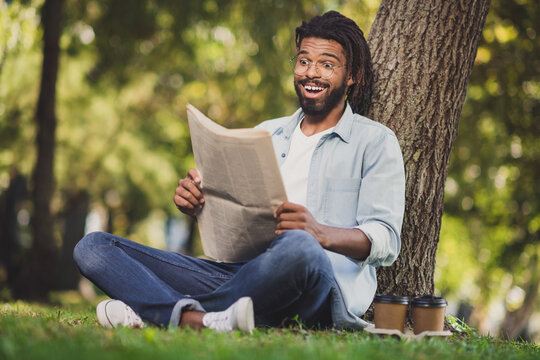 Full size photo of excited brunette afro american man sit grass park read newspaper amazed outside in outdoors weekend