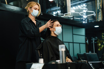 Young woman in face mask doing hairstyle for her client in beauty salon