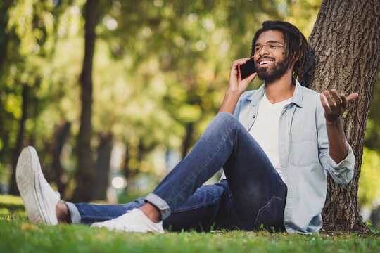 Full length photo of brunette relaxed dark skin man talk phone sit grass in park good mood outside in outdoors