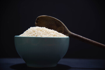 Porcelain pan with raw white rice and wooden spoon on black background.