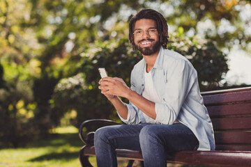 Obraz premium Photo of cheerful positive young afro american man hold phone sit bench park in outside outdoors weekend