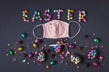 Chocolate Easter eggs full of candies, surrounded by colorful sweets on the grey background. Easter background. Easter sign and pink stylish mask. Easter during coronavirus. Protection against flu