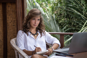 Portrait of young beautiful woman freelancer working with notebook, laptop in white shirt on balcony of tropical bungalow with palm trees view