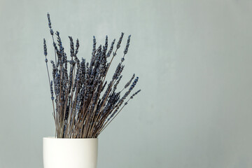 A bouquet of dry blue lavender stands in a white vase on a table against a gray wall. Space for text.