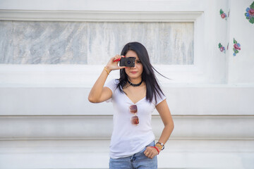 Portrait beautiful Asian woman in a white T-shirt taking a photo directly aimed at the viewer with a white wall in The temple in Thailand