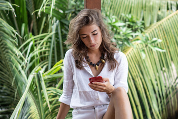Portrait of young beautiful woman freelancer with red smartphone on balcony of tropical bungalow...