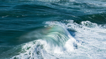 Giant turquoise ocean waves crash on the water surface forming a rainbow