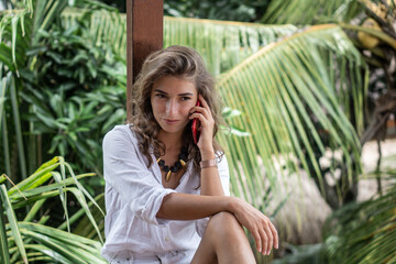 Portrait of young beautiful woman freelancer with red smartphone on balcony of tropical bungalow with palm trees view in Bali