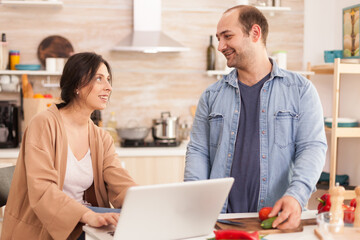 Obraz premium Couple looking at each other while searching recipe on laptop for healthy salad. Man helping woman to prepare healthy organic dinner, cooking together. Romantic cheerful love relationship