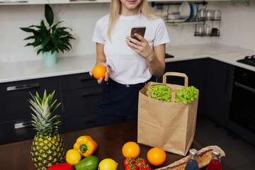Obraz premium Cropped shot of smiling young girl wearing casual clothes using smartphone while near a kitchen table with food purchases.