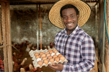 Happy adult male Afro farmer collecting organic eggs in a henhouse - Farm lifestyle and healthy food concept - Focus on face