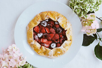 Delicious freshly baked vegan strawberry and cherry galette on white tablecloth background with hydrangea flowers, top view. Sweet food, summer dessert.
