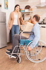 Disabled man in wheelchair with fresh groceries from supermarket and his wife in kitchen. Disabled paralyzed handicapped man with walking disability integrating after an accident.