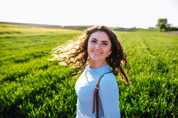 Curly brunette enjoys sunshine in summer day. Portrait of smiling beautiful girl outdoors. Young woman standing on street with curly hair blown by the wind, covering her face.